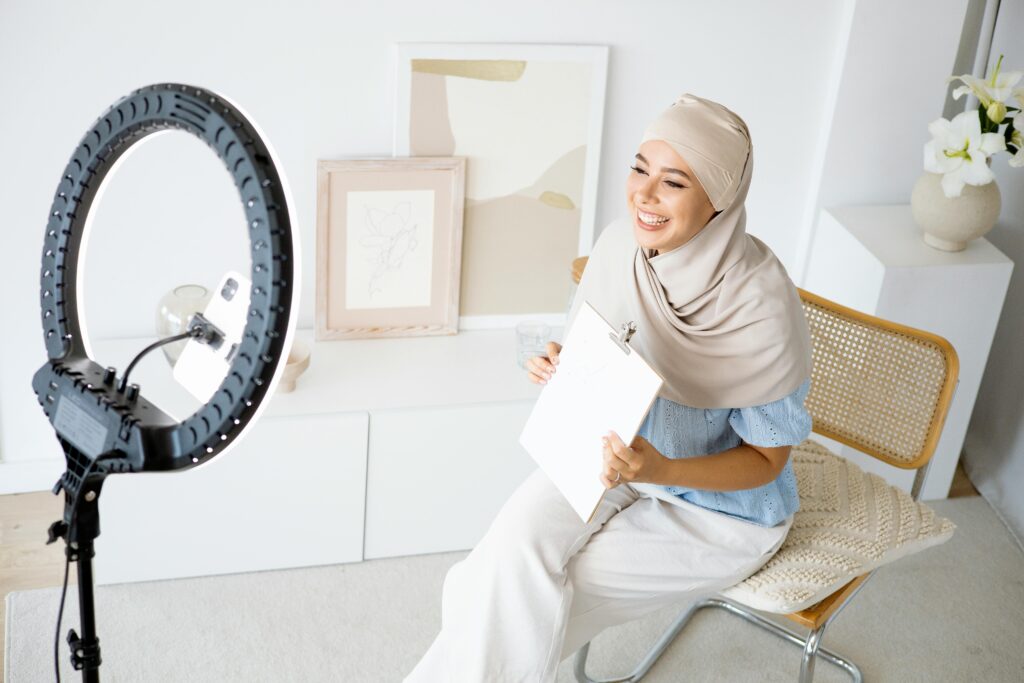 Smiling woman in hijab video blogging using a ring light. Modern online communication setup.